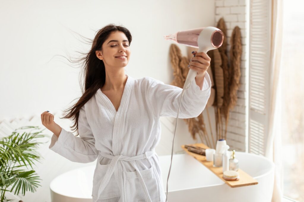 Brunette Woman Drying And Styling Hair With Hairdryer In Bathroom